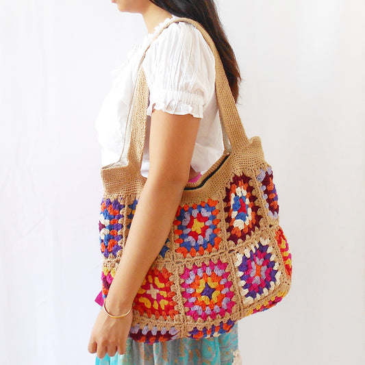 Hand-crocheted tote bag featuring a natural tan/sand colored base and straps, adorned with vibrant, multicolored floral granny squares in shades of pink, orange, blue, purple, and yellow. Model shown from the side carrying the bag against a plain white background. Ethically handcrafted by artisans for Dilmaya Gifts.