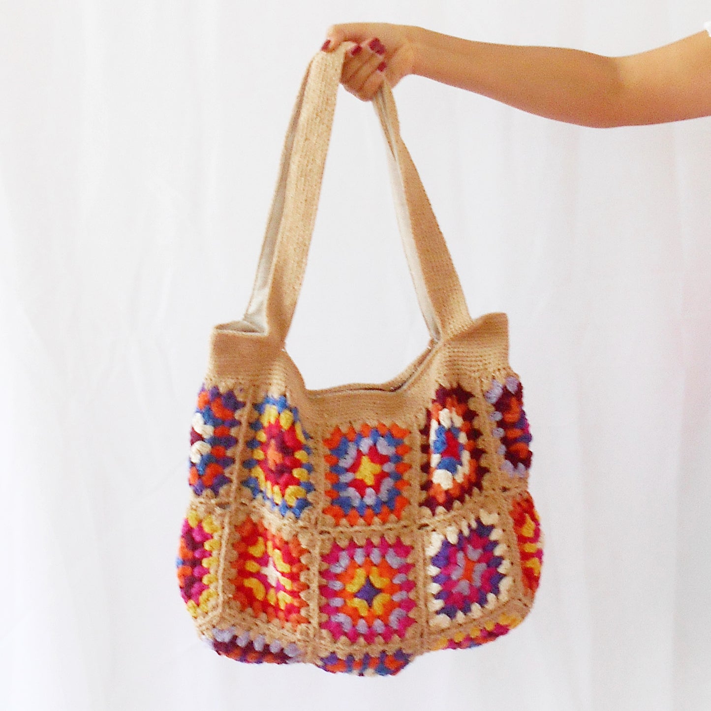 Hand-crocheted tote bag featuring a natural tan/sand colored base and straps, adorned with vibrant, multicolored floral granny squares in shades of pink, orange, blue, purple, and yellow. Model shown from the side carrying the bag against a plain white background. Ethically handcrafted by artisans for Dilmaya Gifts.