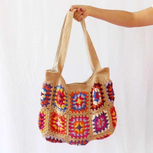 Hand-crocheted tote bag featuring a natural tan/sand colored base and straps, adorned with vibrant, multicolored floral granny squares in shades of pink, orange, blue, purple, and yellow. Model shown from the side carrying the bag against a plain white background. Ethically handcrafted by artisans for Dilmaya Gifts.