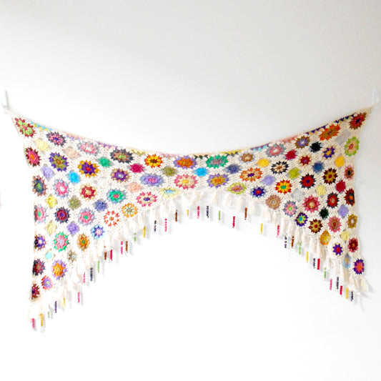 Large hand-crocheted wall hanging made of colorful floral mandala motifs connected by cream yarn, hanging from a corner to create a V-shape against a white wall. Features cream fringe along the edges. Ethically crafted by Nepalese women artisans for Dilmaya Gifts.