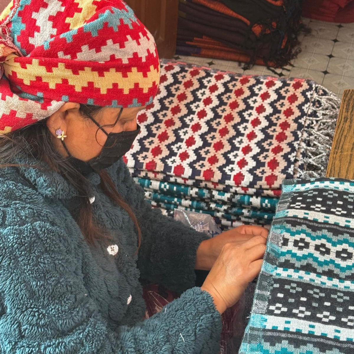 A Nepalese artisan carefully crafting a shawl, surrounded by vibrant woven textiles. She wears a colourful headscarf and a mask while working.