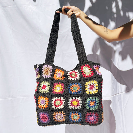 Hand-crocheted Midnight Multi-Floral tote bag featuring colorful granny squares with vibrant flower motifs in red, yellow, orange, blue, pink, and green arranged on a dark background. The shoulder straps are crocheted in matching midnight color. Shown held up against a white wall with dappled shadows.