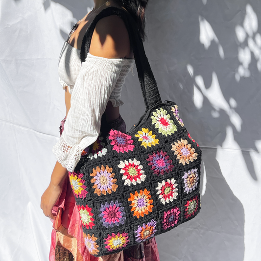 Hand-crocheted Midnight Multi-Floral tote bag featuring colorful granny squares with vibrant flower motifs in red, yellow, orange, blue, pink, and green arranged on a dark background. The shoulder straps are crocheted in matching midnight color. Shown held up against a white wall with dappled shadows.