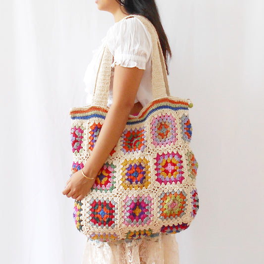 Handcrafted cotton crochet bag in grey-blue with colourful granny squares featuring yellow, orange, pink, and purple patterns, held against white background with natural shadows