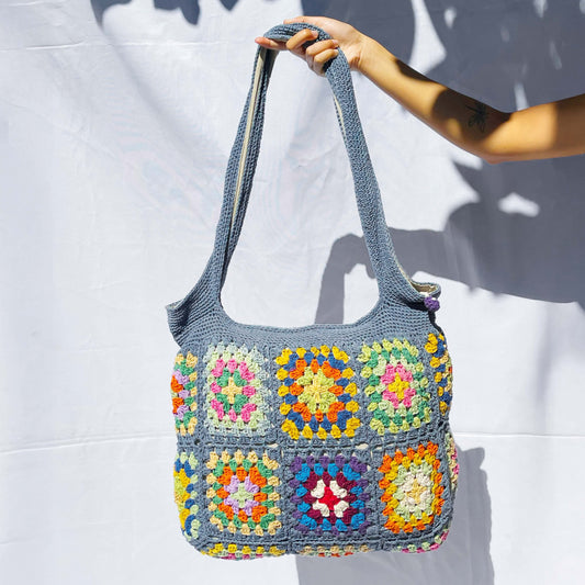 Handcrafted cotton crochet bag in grey-blue with colourful granny squares featuring yellow, orange, pink, and purple patterns, held against white background with natural shadows