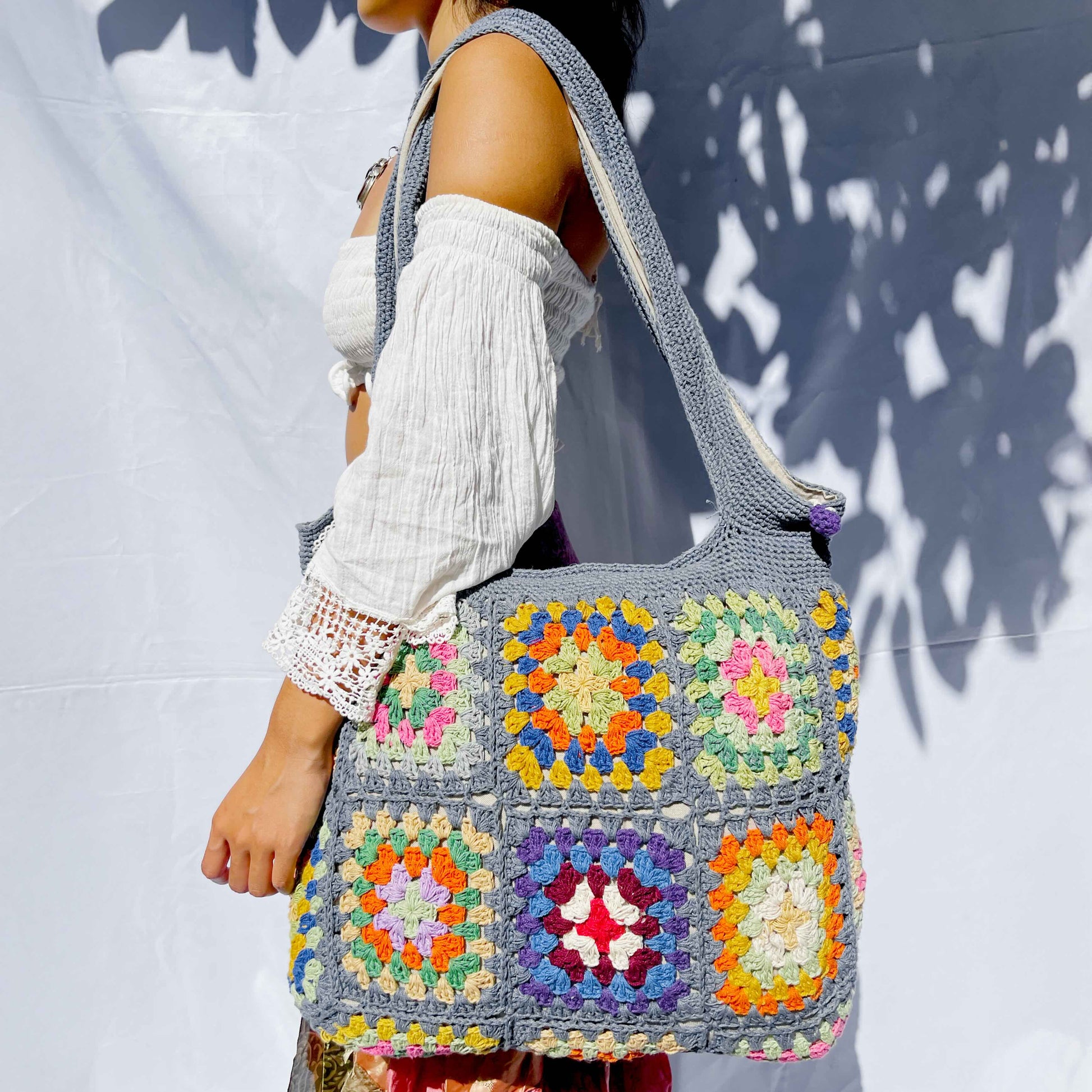 Handcrafted cotton crochet bag in grey-blue with colourful granny squares featuring yellow, orange, pink, and purple floral patterns, held against white background with natural shadows
