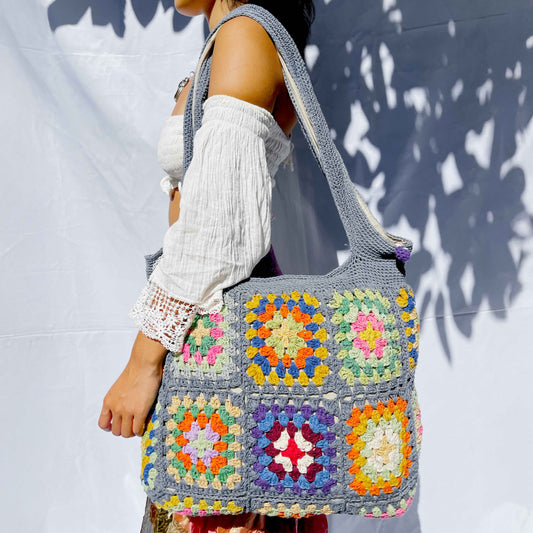 Handcrafted cotton crochet bag in grey-blue with colourful granny squares featuring yellow, orange, pink, and purple floral patterns, held against white background with natural shadows