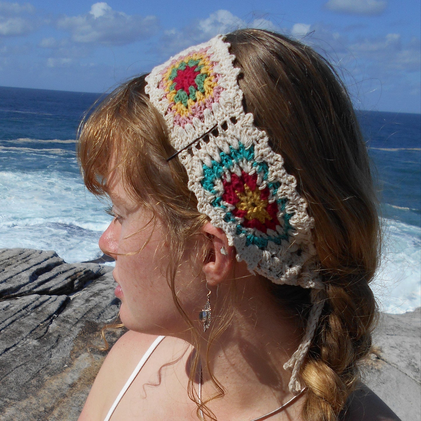 Adjustable hand-crocheted headband featuring cream-colored base with vibrant floral granny squares in teal, red, yellow, and pink. Model with braided hair wearing the headband at the beach, paired with white top and a crystal pendant necklace. Ethically crafted by Nepalese women artisans.