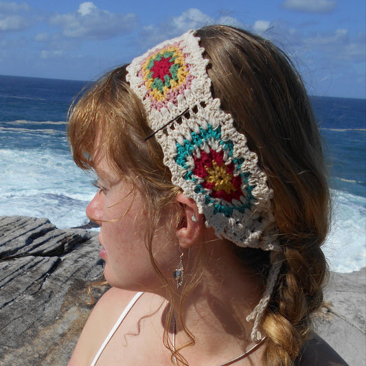 Adjustable hand-crocheted headband featuring cream-colored base with vibrant floral granny squares in teal, red, yellow, and pink. Model with braided hair wearing the headband at the beach, paired with white top and a crystal pendant necklace. Ethically crafted by Nepalese women artisans.