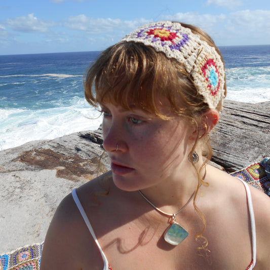 Adjustable hand-crocheted headband featuring cream-colored base with vibrant floral granny squares in teal, red, yellow, and pink. Model with braided hair wearing the headband at beach, paired with white top and crystal pendant necklace. Ethically crafted by Nepalese women artisans .