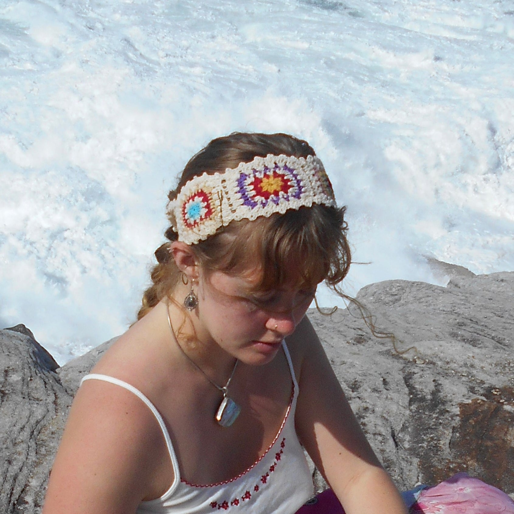 Adjustable hand-crocheted headband featuring cream-colored base with vibrant floral granny squares in teal, red, yellow, and pink. Model with braided hair wearing the headband at beach, paired with white top and crystal pendant necklace. Ethically crafted by Nepalese women artisans.